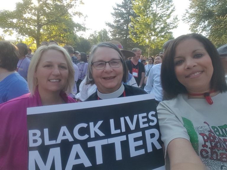 Three women holding a "Black Lives Matter" sign, smile for a selfie.