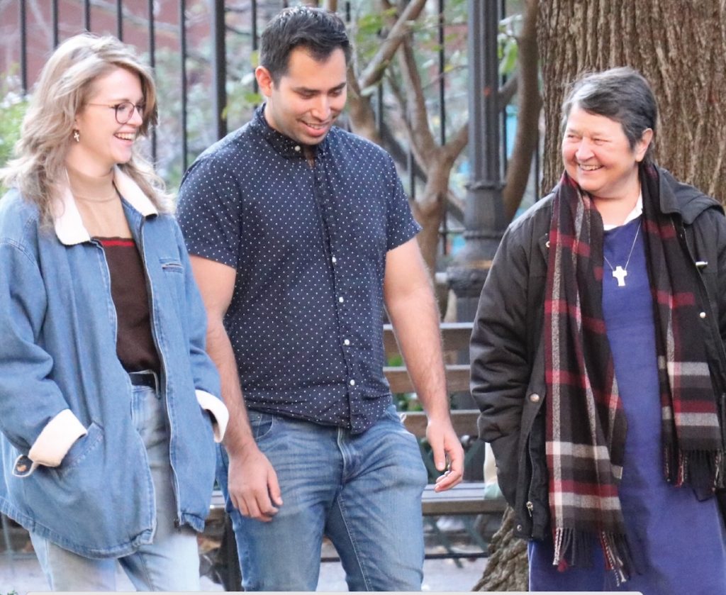 A woman in a purple dress and silver cross around her neck walks with a younger man and woman, all smiling.